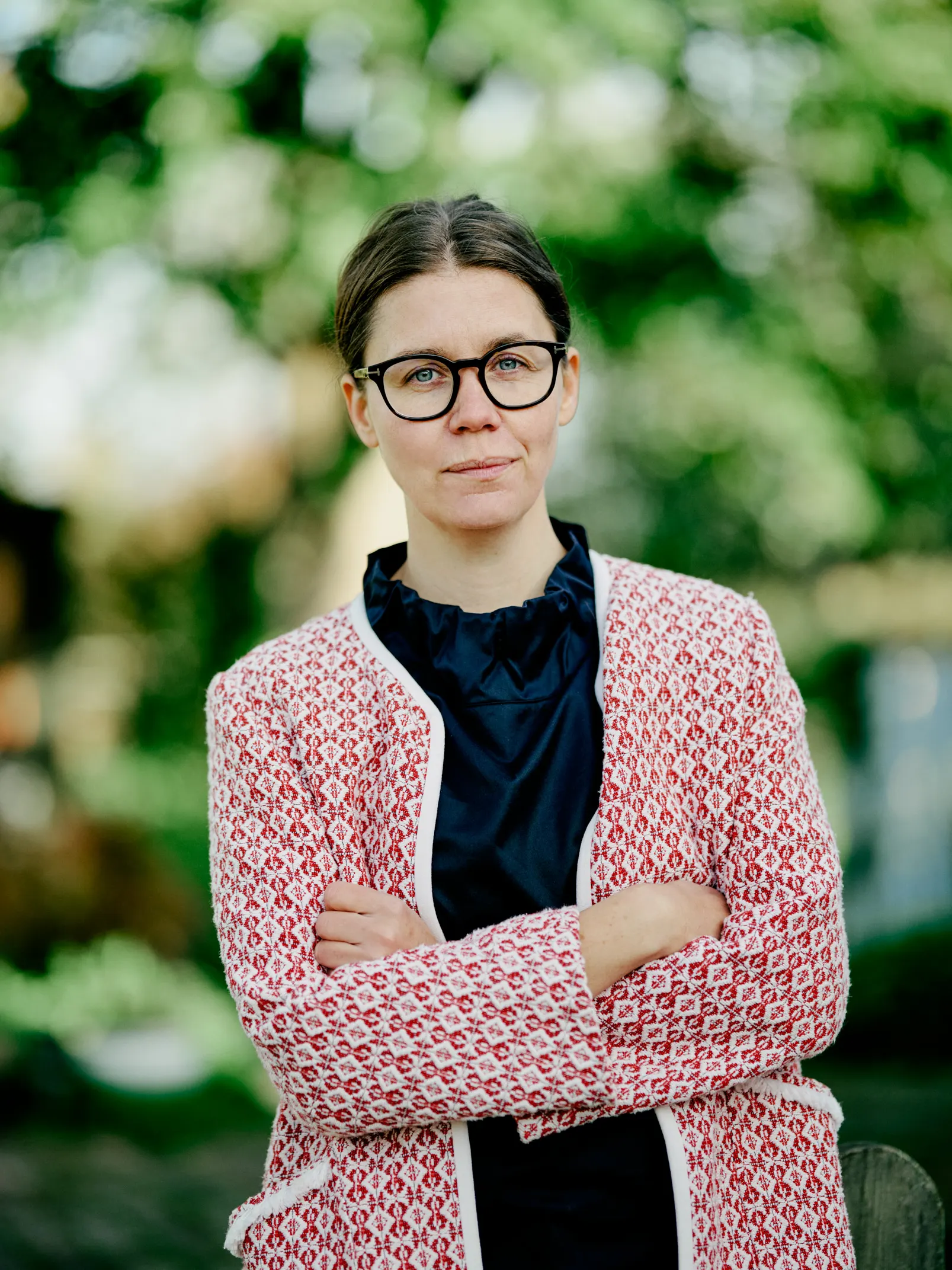 Portrætbillede af en kvindelig advokat med briller, der står med korslagte arme og ser direkte ind i kameraet. Hun bærer en mørk bluse under en rød og hvid mønstret jakke, og baggrunden er en sløret have med grønne træer.